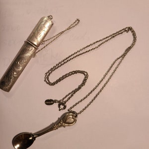 May include: A silver necklace with a heart-shaped spoon pendant and a silver chain. The spoon is engraved with the word "Sterling". The necklace is attached to a silver tube with an engraved design.