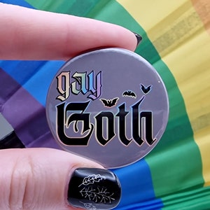 May include: A round button badge with a grey background and the text "gay goth" in black with rainbow-coloured outlines. There are several small black bats around the text.
