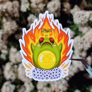 May include: A sticker featuring a green cartoon frog with red eyes and a pentagram on its belly, surrounded by flames. The text "SPAWN OF SATAN" is written on the sticker.
