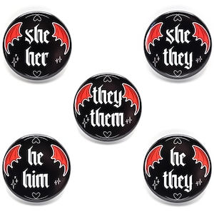 May include: Five black pronoun badges with white gothic lettering and red bat wings. The badges feature the pronouns "she/her", "she/they", "they/them", "he/him", and "he/they". Each badge has heart and star details.