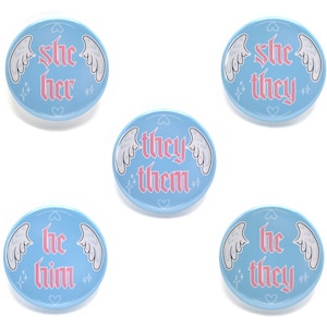 May include: Set of four blue and pink enamel pins with angel wings and text. The pins read "she her", "she they", "they them", and "he him".