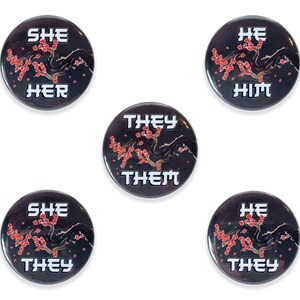 May include: Five round black buttons with a white floral design and white text. The text on the buttons reads "She Her", "He Him", "They Them", "She They", and "He They".