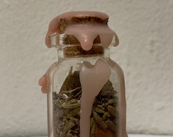 May include: A small, clear glass bottle filled with dried herbs and spices, sealed with a cork and pink wax. The wax drips down the sides of the bottle, creating a decorative effect. The bottle is likely used for storing herbs or potions.
