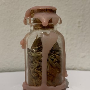 Puede incluir: Una pequeña botella de vidrio transparente llena de hierbas y especias secas, sellada con un corcho y cera rosa. La cera gotea por los lados de la botella, creando un efecto decorativo. La botella probablemente se utiliza para almacenar hierbas o pociones.