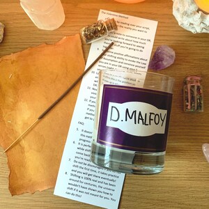 Puede incluir: Un vaso transparente con una etiqueta morada que dice "D.MALFOY" en letras blancas. También se ven un palito de incienso, pequeños frascos y un papel con texto, lo que sugiere un tema espiritual o metafísico.