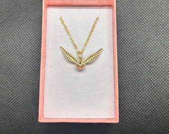 May include: A gold-toned necklace featuring a pendant shaped like a small sphere with two outstretched, textured wings. The necklace is displayed inside a rectangular, light pink jewelry box with a soft, white, felt-like interior lining.