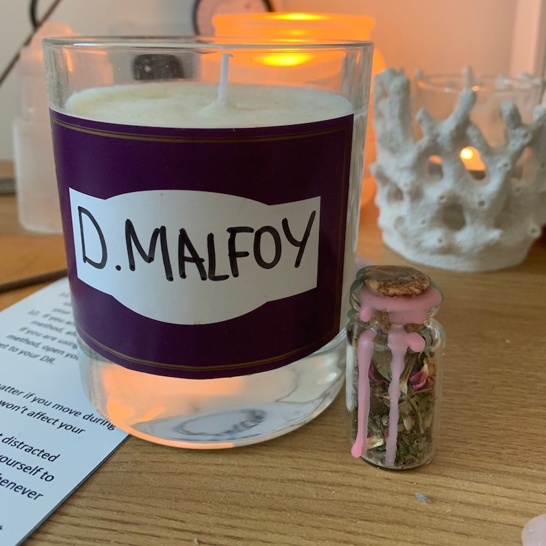 May include: A clear glass candle with a white wax interior and a purple label that reads "D. MALFOY" in white. A small glass bottle with a cork and pink wax is next to the candle. The bottle contains dried herbs and flowers.