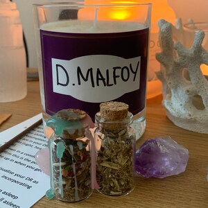 May include: A candle in a glass jar with a purple label that reads "D. MALFOY" in white. Two small glass bottles with cork stoppers sit in front of the candle, along with a purple crystal and a pink crystal.