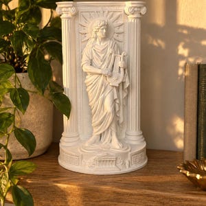 Può includere: Un rilievo scultoreo bianco raffigurante una figura centrale che regge una lira, incorniciata da due colonne classiche. La figura è posizionata davanti a un motivo a raggiera, con una piccola struttura templare raffigurata alla base.