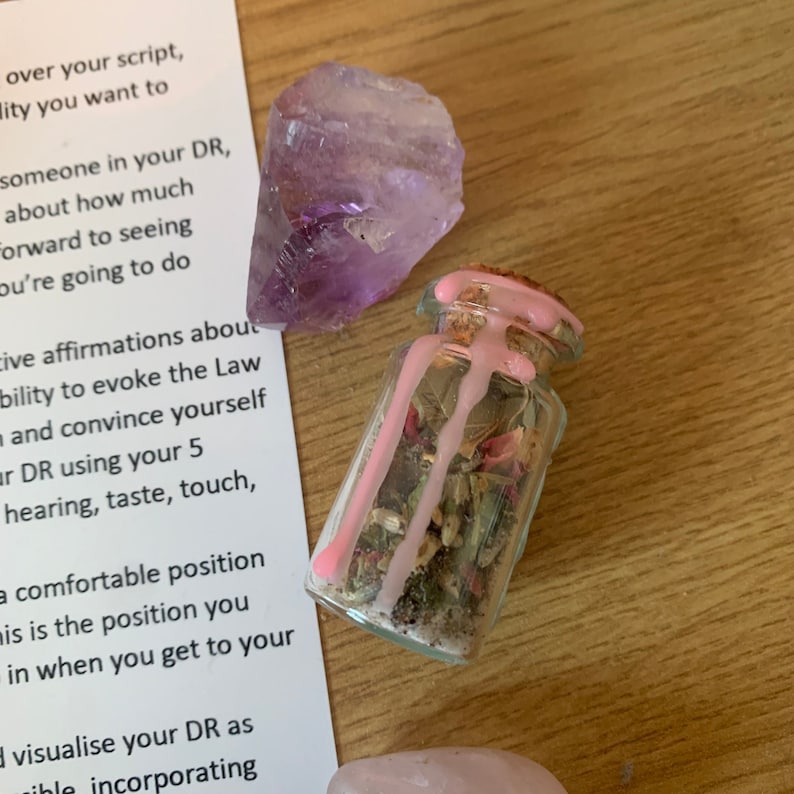 May include: A small glass bottle filled with dried herbs and sealed with pink wax. A large, rough-cut amethyst crystal sits nearby. The items are arranged on a light-colored wooden surface, with a piece of paper with text in the background.