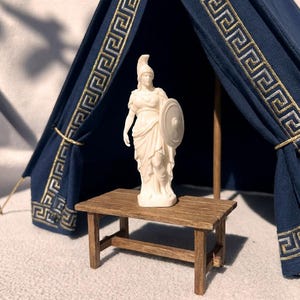 Op de afbeelding: Een wit beeldje van een staand figuur met een helm en een rond schild, geplaatst op een kleine rechthoekige bruine houten tafel. Achter de tafel staat een donkerblauwe stoffen tentstructuur met een goudkleurig Grieks sleutelpatroon.