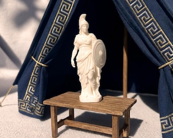 May include: A white figurine of a standing figure wearing a helmet and holding a round shield, positioned on a small brown wooden rectangular table. Behind the table is a dark blue fabric tent structure featuring a gold Greek key pattern.