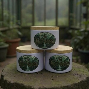 May include: Three white cylindrical containers with wooden lids are stacked on a stone surface. Each container features a green oval design with the text "THE GREENHOUSE" and a waterfall illustration. The background is a greenhouse setting.