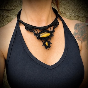 May include: Black macrame necklace featuring a large oval tiger's eye stone and a smaller teardrop-shaped stone. The necklace has intricate woven detailing and a thick, rounded collar. The stones display yellow and brown hues.