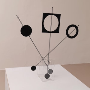 Könnte beinhalten: Abstrakte schwarze Metallskulptur mit geometrischen Formen: ein Kreis, ein Quadrat mit einem Kreis darin und ein Ring. Die Skulptur wird von einem transparenten Acrylständer getragen und schafft ein modernes Kunstwerk.