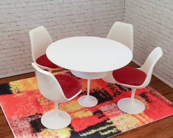 May include: Miniature white table with four matching chairs. The chairs have red seat cushions. The table and chairs are on a colorful rug with red, orange, yellow, and black hues. The background is a white brick wall.