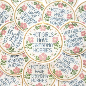 May include: A collection of round stickers with a cross-stitch design. Each sticker features the text "HOT GIRLS HAVE GRANDMA HOBBIES" in blue, surrounded by pink flowers and green leaves, set within a wooden embroidery hoop.