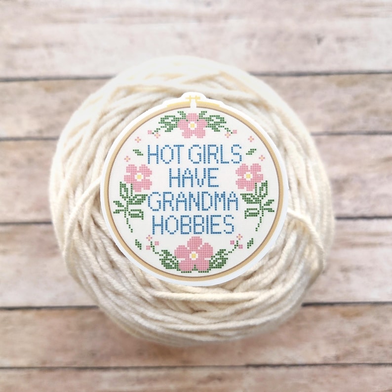 May include: A round, off-white sticker with the text "HOT GIRLS HAVE GRANDMA HOBBIES" in blue cross-stitch style lettering, surrounded by pink flowers and green leaves. The sticker is set against a ball of cream-colored yarn.