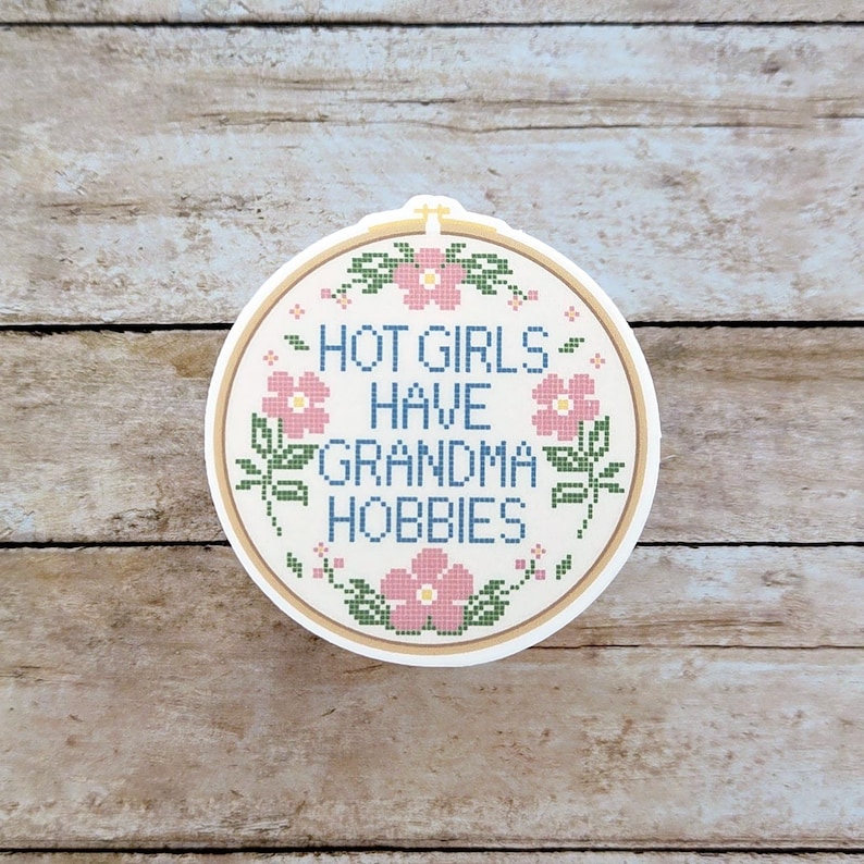 May include: A round sticker with a cross-stitch design. The sticker features the text "HOT GIRLS HAVE GRANDMA HOBBIES" in blue, pink, and green. The design is set within a beige embroidery hoop.