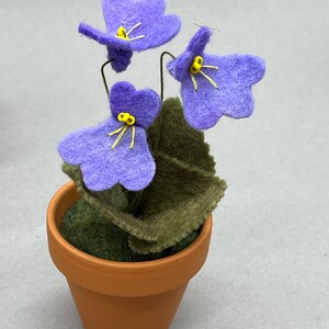 以下が含まれることがあります： 小さなテラコッタの鉢に植えられた、黄色い中心と緑の葉を持つ3つの紫色の花が特徴の手作りのフェルトフラワーアレンジメント。 花は柔らかいフェルトで作られ、繊細なワイヤーの茎が付いています。