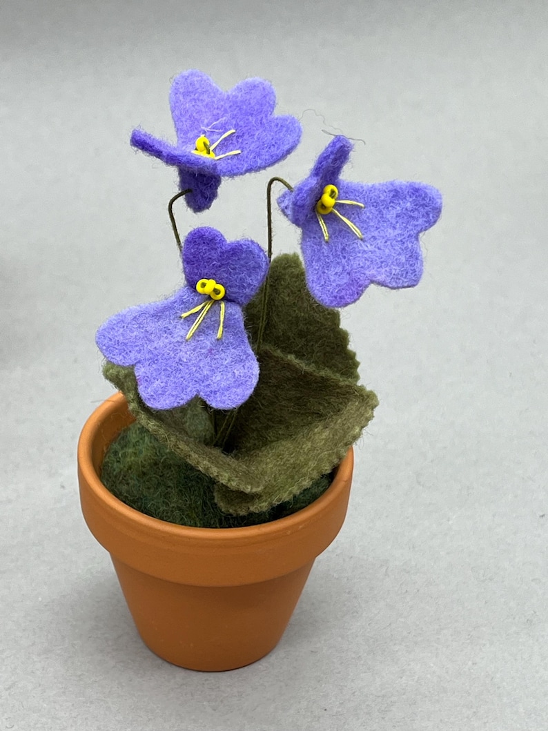 以下が含まれることがあります： 小さなテラコッタの鉢に植えられた、黄色い中心と緑の葉を持つ3つの紫色の花が特徴の手作りのフェルトフラワーアレンジメント。 花は柔らかいフェルトで作られ、繊細なワイヤーの茎が付いています。