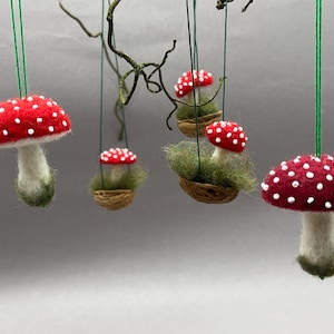 Puede incluir: Adornos de setas de fieltro hechos a mano con sombreros rojos y puntos blancos, suspendidos por una cuerda verde. Algunas setas están en pequeñas cestas tejidas con musgo verde. Una decoración caprichosa inspirada en la naturaleza.