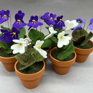 以下が含まれることがあります： 小さなテラコッタの鉢植えのコレクション。それぞれにフェルトの花のアレンジメントが入っています。花は濃い紫または白で、黄色の中心と緑の葉があります。鉢は緑の苔で満たされています。