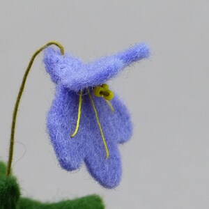 以下が含まれることがあります： 手作りの水色のフェルトの花のクローズアップ。黄色の雄しべと細い茶色の茎があります。花は質感があり、無地の明るい灰色の背景に配置されています。花びらは柔らかく丸みを帯びています。