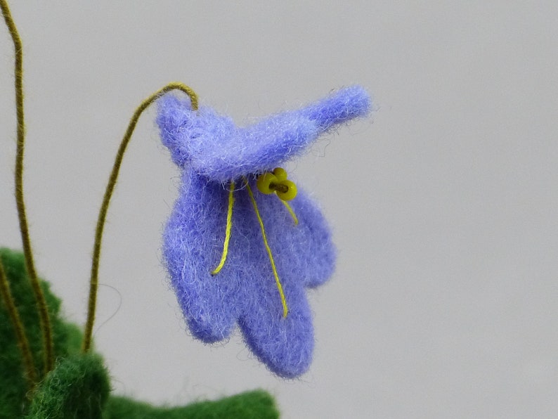 以下が含まれることがあります： 手作りの水色のフェルトの花のクローズアップ。黄色の雄しべと細い茶色の茎があります。花は質感があり、無地の明るい灰色の背景に配置されています。花びらは柔らかく丸みを帯びています。