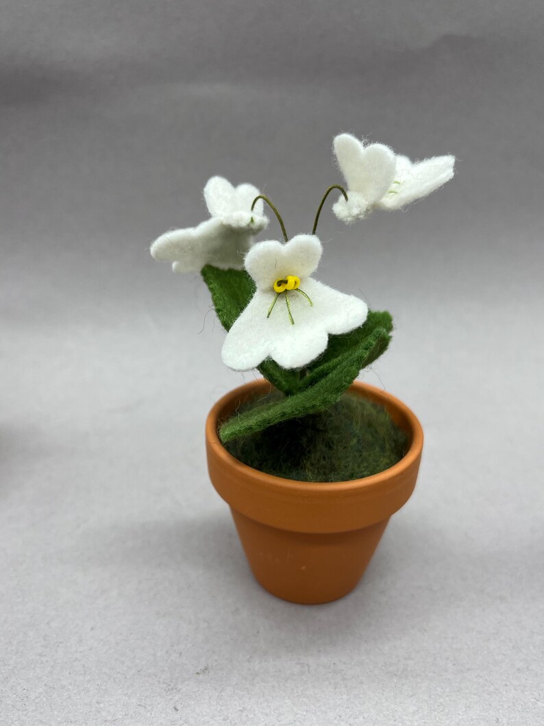以下が含まれることがあります： 小さなテラコッタの鉢植えに飾られたフェルトフラワーアレンジメント。黄色い中心と緑の葉を持つ3つの白いフェルトの花が特徴です。鉢は緑の苔で満たされています。