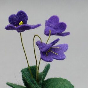 以下が含まれることがあります： 紫色の花びらと黄色の中心を持つフェルトの花のクローズアップ。花には緑色の茎と葉があります。背景は無地の明るいグレーです。