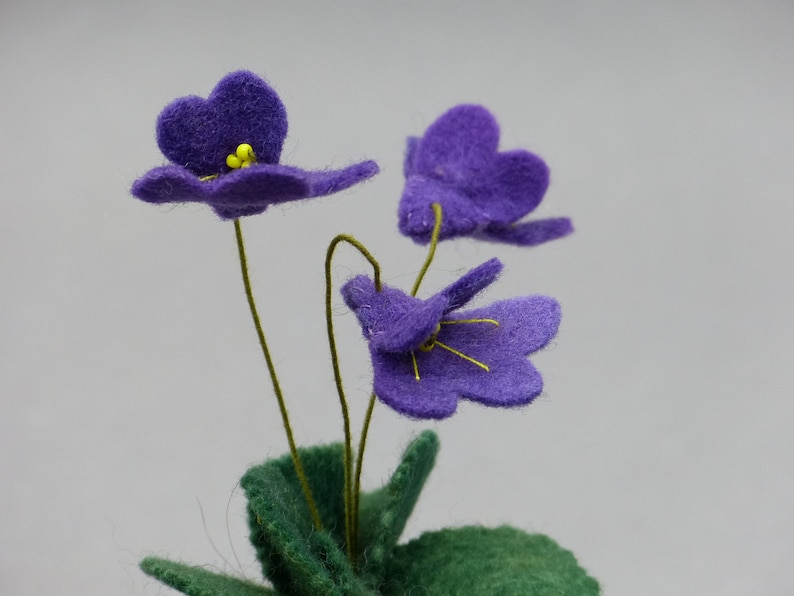 以下が含まれることがあります： 紫色の花びらと黄色の中心を持つフェルトの花のクローズアップ。花には緑色の茎と葉があります。背景は無地の明るいグレーです。