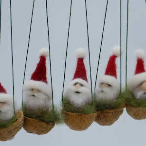 Op de afbeelding: Vijf vilten kerstman ornamenten die aan touwtjes hangen. Elke kerstman draagt een rode muts en een witte baard en zit in een walnootschelp.
