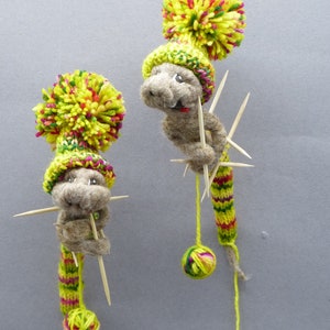 Puede incluir: Dos criaturas caprichosas hechas a mano con fieltro, con sombreros de pompón de hilo y cuerpos de punto. Cada figura tiene brochetas de madera y una pequeña bola de hilo adjunta. Los sombreros son amarillos con detalles rojos y verdes, y los cuerpos son verdes y amarillos.