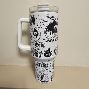 May include: White insulated travel mug with a black Halloween design featuring bats, pumpkins, and characters from a popular animated film. The mug has a white handle and a clear lid. The mug is approximately 25 cm tall.
