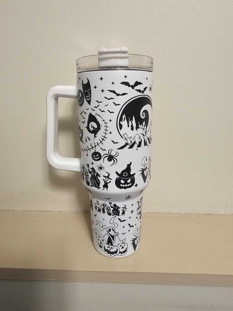 May include: White insulated travel mug with a black Halloween design featuring bats, pumpkins, and characters from a popular animated film. The mug has a white handle and a clear lid. The mug is approximately 25 cm tall.