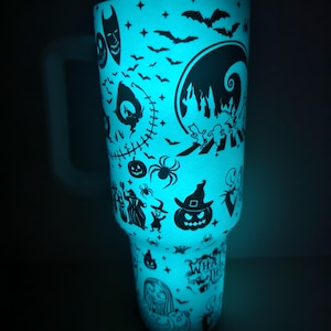 May include: A teal-coloured tumbler with a glowing effect, decorated with black silhouettes of Halloween characters. The design includes bats, Jack Skellington, Sally, and other characters from The Nightmare Before Christmas. It has a white handle.