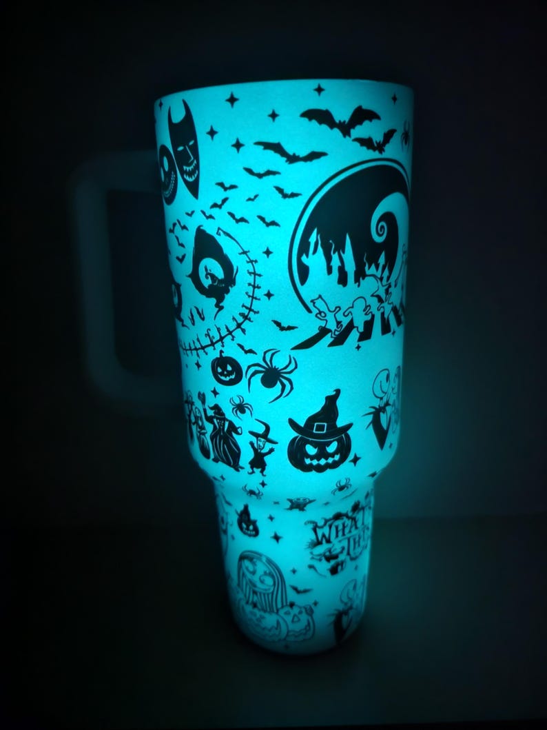 May include: A teal-coloured tumbler with a glowing effect, decorated with black silhouettes of Halloween characters. The design includes bats, Jack Skellington, Sally, and other characters from The Nightmare Before Christmas. It has a white handle.