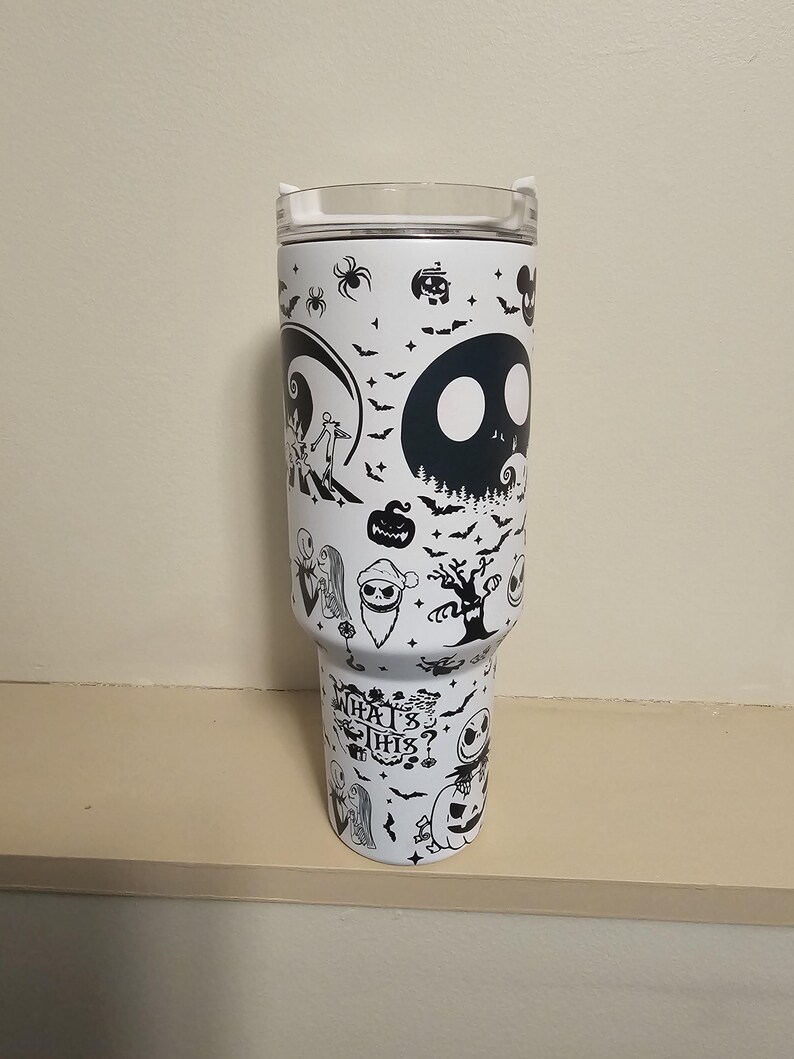 May include: White insulated tumbler with black illustrations of characters and scenes from The Nightmare Before Christmas. The tumbler has a clear lid and the words "What's This?" in a stylized font.