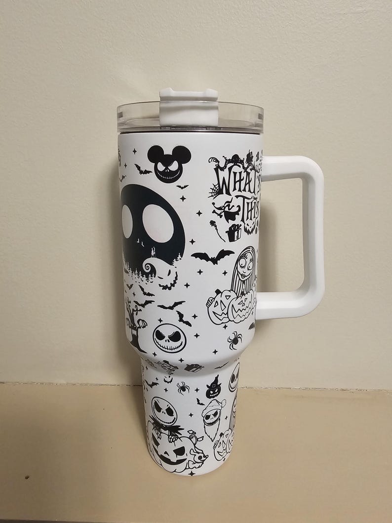May include: White travel mug with a clear lid and white handle. The mug is decorated with black illustrations of characters and imagery from "The Nightmare Before Christmas", including Jack Skellington and the text "What's This?"