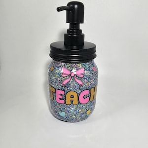 May include: A clear glass jar with a black pump dispenser and a glittery blue, green, and pink design. The jar has the word "TEACH" written on it in pink and gold letters.
