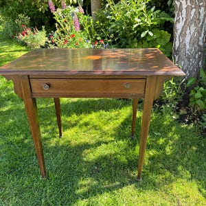 May include: A rectangular wooden side table featuring a smooth, polished brown surface with a central inlay design. The table includes a single horizontal drawer equipped with two small, round knobs and is supported by four slender, tapered legs.