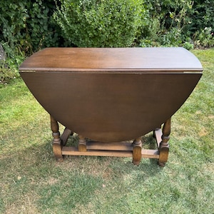 May include: A dark brown wooden drop-leaf table featuring a rectangular top with rounded edges and two hinged side leaves that are currently folded down. The table is supported by four turned legs connected by a sturdy rectangular base frame.