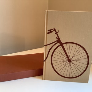 Puede incluir: Dos libros de tapa dura con cubiertas marrones y beige. El libro de la derecha tiene una silueta de una bicicleta con una rueda delantera grande en la portada.