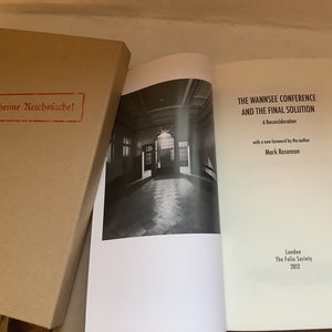 May include: A book titled "THE WANNSEE CONFERENCE AND THE FINAL SOLUTION" with a black and white photo of a hallway. The book is open next to a tan box with the text "Geheime Reichssache!" stamped in red.