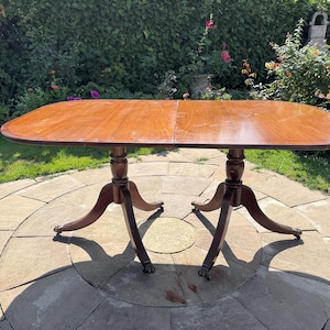 May include: A rectangular wooden dining table with rounded corners and a smooth, polished brown finish. The table is supported by two central pedestals, each featuring four curved legs that terminate in small, detailed claw-style feet.