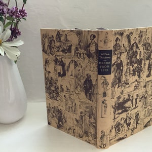 Op de afbeelding: Een vintage boek met een bruine omslag met een herhalend patroon van zwart-wit illustraties. De titel van het boek is "William Thackeray's Drawn From Life".