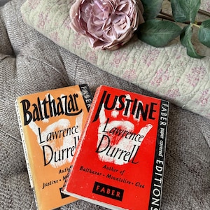 May include: Two paperback books by Lawrence Durrell titled Balthazar and Justine, featuring orange and red covers with a white handprint graphic, black text, and the Faber publisher logo, positioned near a large, pale pink artificial flower.