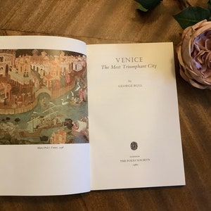 May include: A book titled "Venice: The Most Triumphant City" by George Bull. The book cover features a painting of a cityscape with a boat in the water. The book was published by The Folio Society in 1980.