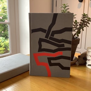 Op de afbeelding: Een grijs hardback boek met een zwart en rood abstract ontwerp op de omslag. Het ontwerp bevat een reeks van elkaar kruisende lijnen en vormen.